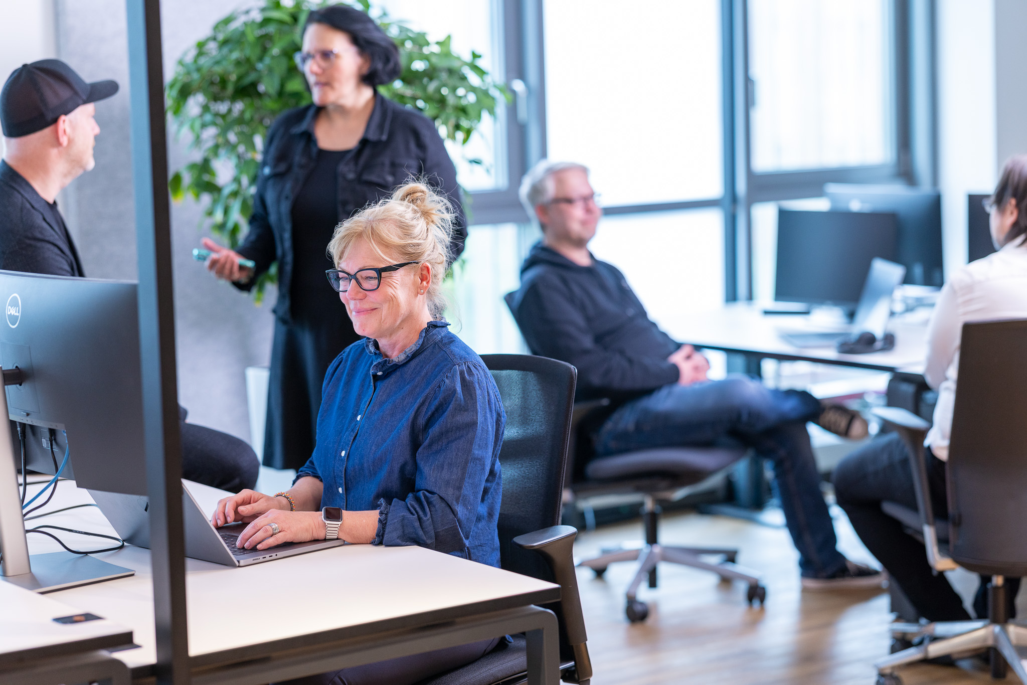 Woman working on a laptop, colleagues in the office behind her